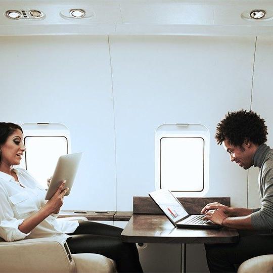 一男一女坐在一架私人飞机的桌子旁，用笔记本电脑和平板电脑与商务航空连接