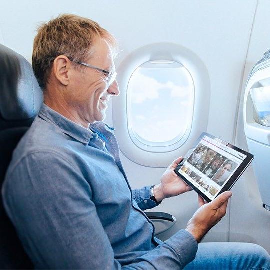 A smiling man wearing glasses is holding a tablet viewing inflight marketing content on his tablet