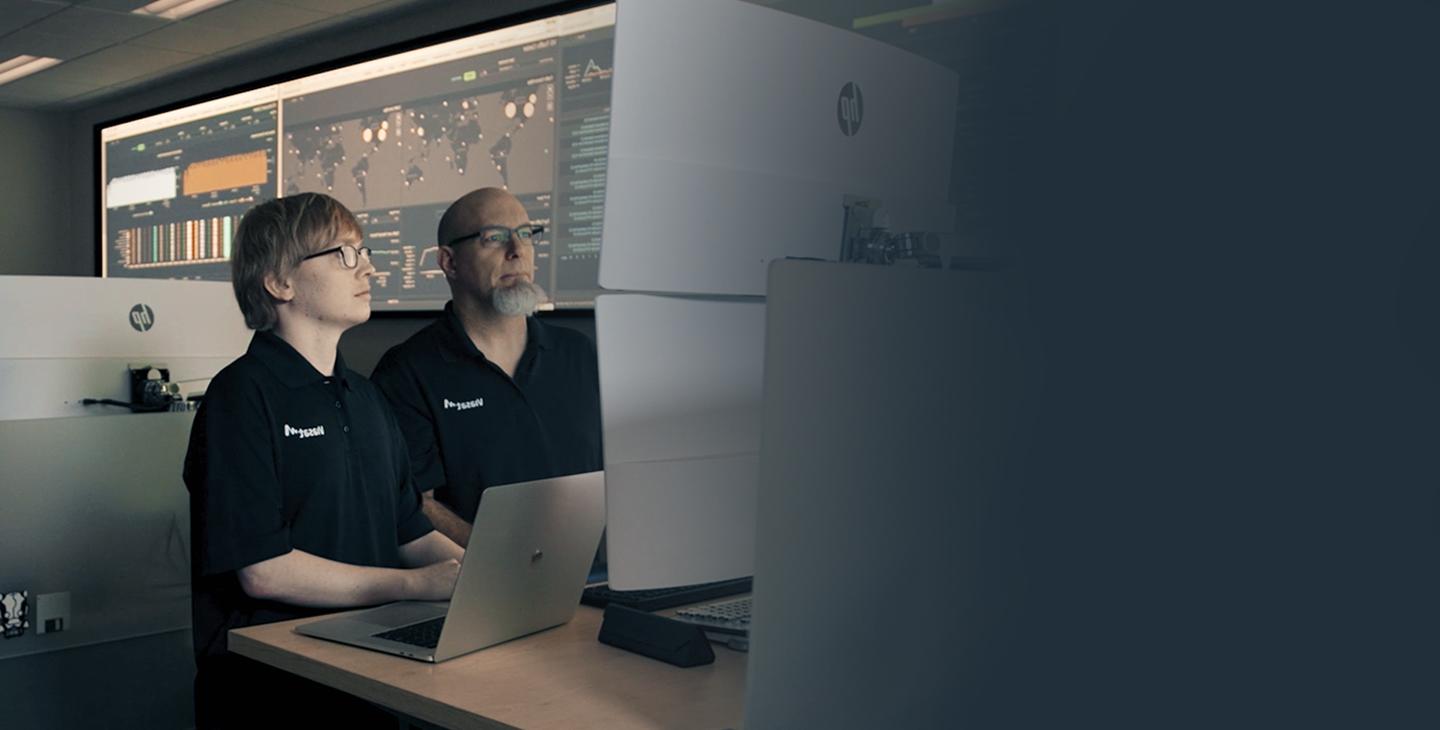 两名Viasat的男性员工穿着带有白色Viasat标志的黑色polo衫，在一家网络安全运营中心工作